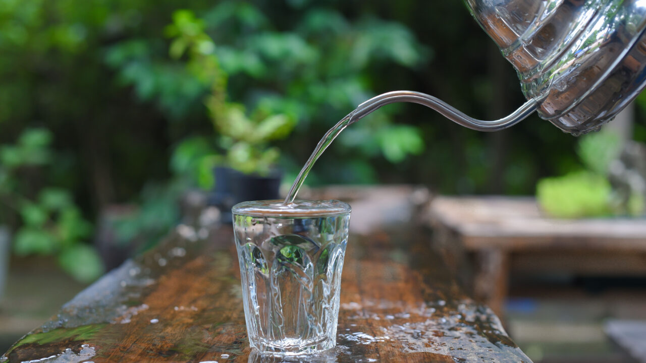 Drinking water poured in glass on table from silver kettle.