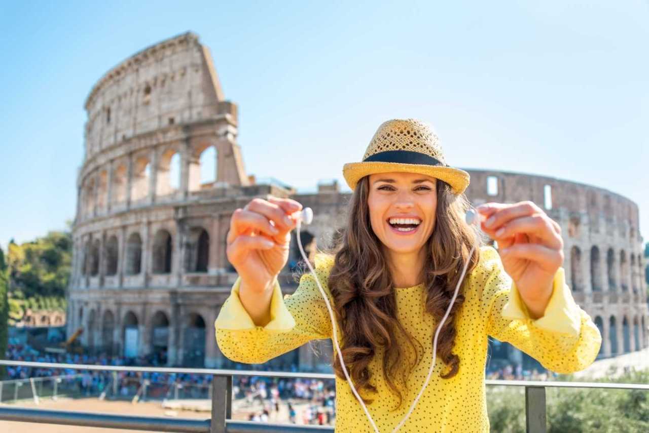 Happy young woman showing headphones with audio guide in front of colleseum in Rome.