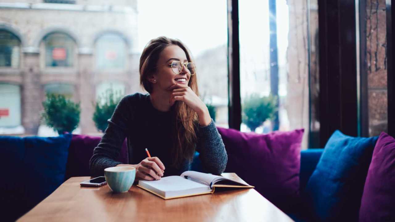 Positive woman at cafe table writing in journal with cup of coffee.