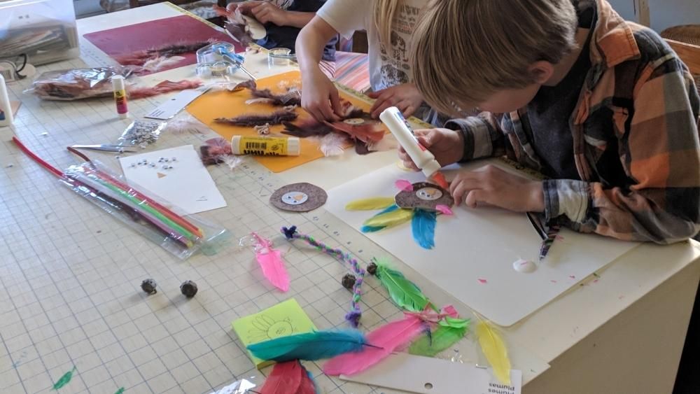 Kids sit at a table doing crafts with many craft supplies spread out. They're gluing feathers on googley eyed turkey crafts