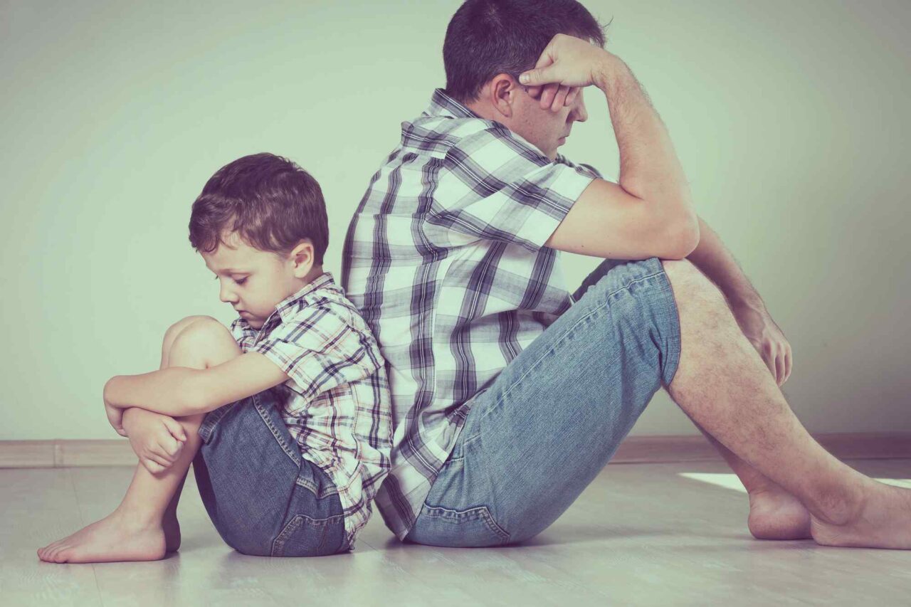 Sad son and his dad sitting on the floor with backs to one another looking upset.